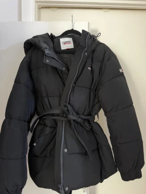Svart pufferjacka Tommy Hilfiger - Snygg svart pufferjacka från Tommy Hilfiger med huva och midjebälte för en schysst siluett. Jackan har dragkedja, tryckknappar och små logodetaljer på bröstet och ärmen. Perfekt för kalla dagar när du vill hålla stilen.