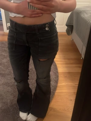 Svarta bootcut jeans med slitningar - Säljer ett par svarta bootcut jeans med slitningar på knäna och dubbla knappar i midjan. Jeansen har hög midja och raka ben som går ut lite nedtill. Snygga detaljer och trendig passform, perfekta till en avslappnad stil.