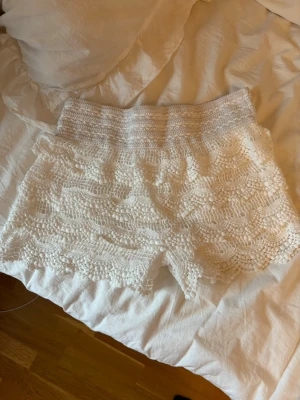 Vita virkade shorts med spetsdetaljer - Säljer ett par vita virkade shorts med fina spetsdetaljer och vågig kant. Shortsen har elastisk midja och är perfekta för sommaren. De är lätta och luftiga, vilket gör dem riktigt sköna under varma dagar. Passar xs-m då dem är strshiha 