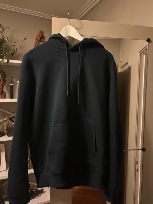 Mörkblå hoodie med ficka - En stilren mörkblå hoodie med huva och dragsnören. Hoodien har en stor magficka framtill och ribbade muddar vid ärmslut och nederkant. Perfekt för en avslappnad look och enkel att matcha med jeans eller joggers.