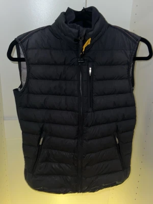 Parajumpers dunväst Y-M (barn M) svart - Snygg svart dunväst från Parajumpers med klassisk gul detalj vid kragen. Västen har dragkedja framtill, två sidofickor med dragkedja och en bröstficka. Quiltad design och hög krage för extra värme. Perfekt för lager-på-lager-stil. Finns små fläckar längst ner men märks inte om inte man kollar jätte noga finns små håll fram och back men annars top skick