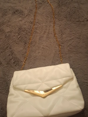 Vit quiltad axelväska med guldkedja - Snygg vit axelväska i quiltat mönster med guldfärgad kedja och dekorativt guldfärgat spänne framtill. Väskan har en mjuk, vadderad känsla och modern design som passar till många olika outfits. Perfekt för dig som vill ha en trendig accessoar.