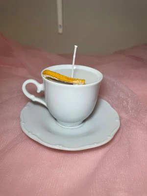 Doftljus i vaniljdoft i en vit tekopp med fat - Det här ljuset är gjort av återanvändbara material. Tekoppen är köpt secondhand och stearinet är från kyrkor som tänkte slänga det. Det här ljuset har en svag doft utav vanilj, så känsliga även kan använda det. Det här ljuset har även torkade blommor och apelsin på som dekoration. 