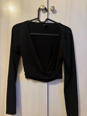 Svart croppad omlottopp - Snygg svart croppad omlottopp med djup v-ringning och långa ärmar. Perfekt för att styla med höga jeans eller kjol. Tunn och stretchig kvalitet som sitter bekvämt.