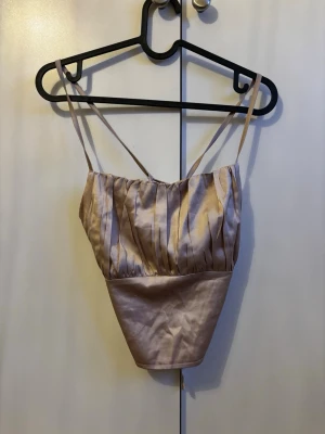 Champagnefärgad satinlinnetopp - Snygg champagnefärgad linnetopp i satin med tunna axelband och öppen rygg med snörning. Toppen har en croppad passform och ger en elegant look med sitt glansiga material. Perfekt att styla med höga jeans eller kjol.