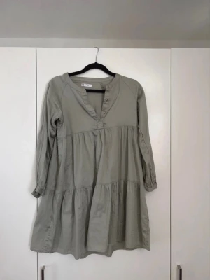 Ljusgrön volangklänning med knappar - Ljusgrön klänning med volanger och v-ringning med knappar framtill. Långärmad modell med avslappnad passform och resår vid ärmslut. Tillverkad i ett mjukt, vävt material som ger en luftig känsla.