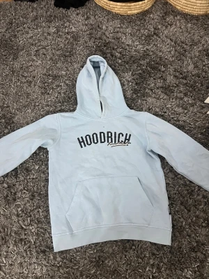 Ljusblå hoodie från Hoodrich - Snygg ljusblå hoodie från Hoodrich med svart tryck på bröstet. Klassisk känguruficka framtill och stor huva. Perfekt för en avslappnad streetwear-look. Tillverkad i mjukt bomullsmaterial som känns skönt mot huden.