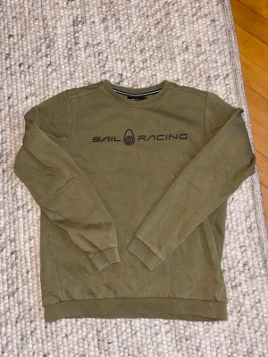 Olivgrön sweatshirt från Sail Racing - Olivgrön crewneck sweatshirt från Sail Racing med svart logotyptryck på bröstet och en mindre logga i nacken. Tröjan har rund hals, långa ärmar och ribbade muddar vid ärmslut och nederkant. Perfekt för en avslappnad och sportig stil.
