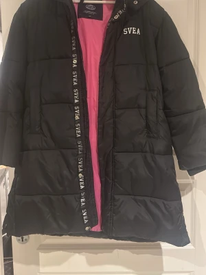 Svart lång dunjacka från SVEA - Svart lång dunjacka från SVEA med huva och dragkedja. Nypris är 1700kr! Jackan har rosa foder, stora fickor och SVEA-logga både fram och bak samt på dragkedjan. Perfekt för kalla vinterdagar och riktigt snygg streetstil.