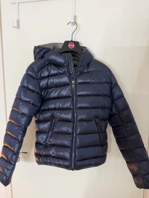 Mörkblå Colmar pufferjacka med huva - Säljer en mörkblå pufferjacka från Colmar med glansig finish och huva. Jackan har dragkedja framtill, två fickor med dragkedja och Colmar-logga på ärmen och nere på baksidan. Colmar storlek 40 motsvarar svensk storlek 34. Köpt i 2024 för cirka 5000 och har använt flitigt men är i väldigt bra skick. 