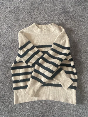 Beige stickad tröja med svarta ränder - Stickad tröja från H&M i beige med breda svarta horisontella ränder. Tröjan har rund halsringning, långa ärmar och ribbade muddar. Perfekt för dig som gillar klassisk och avslappnad stil.