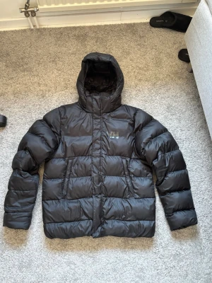 Svart pufferjacka från Helly Hansen - Säljer en svart pufferjacka från Helly Hansen med huva och diskret logga på bröstet. Jackan har breda, vadderade paneler och dragkedja framtill. Perfekt för kalla vinterdagar och riktigt skön att bära.