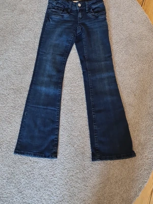 Mörkblå bootcut jeans - Snygga mörkblå bootcut jeans med klassisk femficksdesign och knappgylf. Jeansen har en lätt tvättad look och vida ben som ger en retrovibe. Perfekta att matcha med sneakers eller boots för en avslappnad stil.