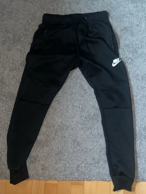 Svarta Nike mjukisbyxor med logga - Säljer ett par svarta mjukisbyxor från Nike med vit logga på vänster ben. Byxorna har dragsko i midjan, muddar vid bensluten och dragkedjefickor på sidorna. Perfekta för chill eller träning, riktigt sköna och stilrena.