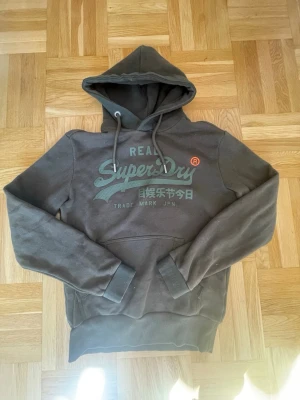 Superdry hoodie - Snygg m hoodie från Superdry med stor logga och japansk text på bröstet. Klassisk känguruficka framtill och justerbar huva med dragsko. Tillverkad i mjukt bomullsmaterial som är skönt mot huden. Perfekt för en avslappnad streetwear-look. 