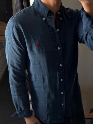 Blå linneskjorta från Ralph Lauren - Klassisk blå linneskjorta från Ralph Lauren med knappslå och vit knappdetalj. Skjortan har lång ärm och ikoniska röda logotypen broderad på bröstet. Tillverkad av 100% linne för en bekväm känsla och avslappnad passform. Den är ganska urtvättad därav priset, annars bra skick