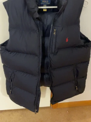 Mörkblå dunväst från Polo Ralph Lauren - Snygg mörkblå dunväst från Polo Ralph Lauren med hög krage, två dragkedjefickor och en bröstficka. Klassisk röd logga på bröstet och quiltad design. Perfekt för lager på lager och håller dig varm under kyliga dagar.