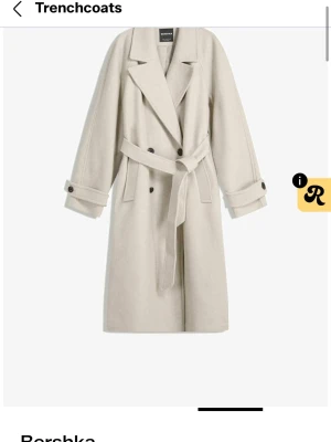 Beige trenchcoat från Bershka - Stilren beige trenchcoat från Bershka med klassisk dubbelknäppning, bred krage och knytskärp i midjan. Jackan har långa ärmar med dekorativa knappar vid ärmsluten och två sidofickor. Perfekt för en trendig och tidlös look.