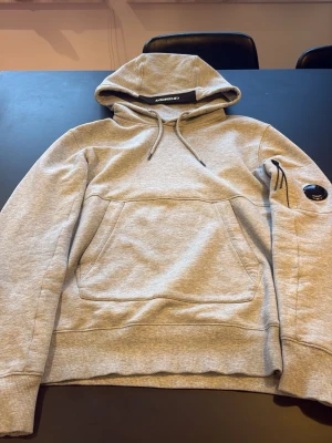 Grå hoodie från C.P. Company - Snygg grå hoodie från C.P. Company med klassisk huva och dragsko. Hoodien har en stor magficka och den ikoniska linsen på vänster ärm. Tillverkad i mjuk bomullsblandning som känns skön mot huden. Perfekt för en avslappnad streetwear-look. Storlek xs/s köparen står för frakt hör av er vid frågor eller bilder 
