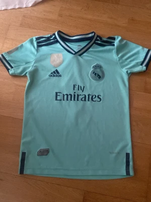 Real Madrid turkos matchtröja Adidas 2019 - Snygg turkos Real Madrid matchtröja från Adidas med mörkblå detaljer och klubbmärke på bröstet. Tröjan har V-ringning, korta ärmar och Fly Emirates-tryck på framsidan. Perfekt för fotbollsträning eller att bära till vardags.