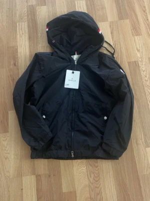 Svart vindjacka från Monclerr - Snygg svart vindjacka från Moncler med huva och dragkedja framtill. Jackan har två fickor med tryckknappar, elastisk nederkant och en Moncler-logga på ärmen. Materialet är lätt och vattenavvisande, perfekt för blåsiga dagar.