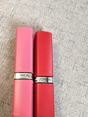 Två L'Oréal Paris läppstift rosa/röd - Säljer två snygga läppstift från L'Oréal Paris, ett i en mjuk rosa nyans och ett i klassiskt rött. Hylsorna är i plast med metalliskt band och har en stilren, modern design. Perfekta för dig som vill ha både färg och elegans i sminkväskan.