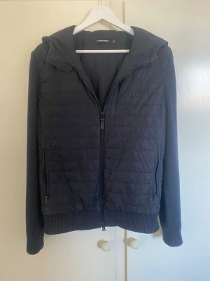 Svart cardigan från J.Lindeberg - Svart cardigan jacka från J.Lindeberg med quiltad framsida och rygg, stickade ärmar och ribbade muddar. Jackan har huva, dragkedja framtill och två sidofickor med dragkedja. Materialmix med både vadderad och stickad känsla för en modern look. Det finns ett litet hål i insidan av ärmen som varken känns eller syns alls när den används. Om fler bilder önskas är det bara att höra av sig.