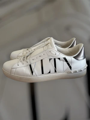 Valentino open inga defekter! - Snygga vita sneakers från Valentino med stor svart VLTN-logga på sidan. Skorna har silvriga detaljer vid hälen och dekorativa nitar längs sulan. Tillverkade i slätt läder med klassisk låg profil och vita snören. Perfekta för dig som gillar exklusiv streetstyle.