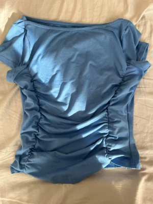 Blå ribbad topp med rynkade sidor - Snygg blå topp med korta ärmar och rynkade detaljer längs sidorna som ger en cool effekt. Tillverkad i mjuk bomull och har en figurnära passform. Perfekt att styla med jeans eller kjol för en trendig look.