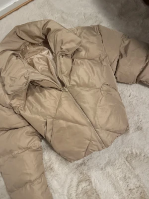 Beige pufferjacka med hög krage - Säljer en oanvänd beige pufferjacka med hög krage och dragkedja framtill. Jackan har en boxig passform och breda, vadderade kanaler som ger en riktigt fluffig look. Perfekt för kalla vinterdagar och enkel att matcha till olika outfits.