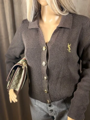 Mörkbrun YSL cardigan med guldknappar - Snygg mörkbrun cardigan från Yves Saint Laurent med ribbad struktur och guldfärgade knappar framtill. Klassisk krage och YSL-logga broderad i gult på bröstet. Perfekt för dig som vill ha en lyxig och stilren look.