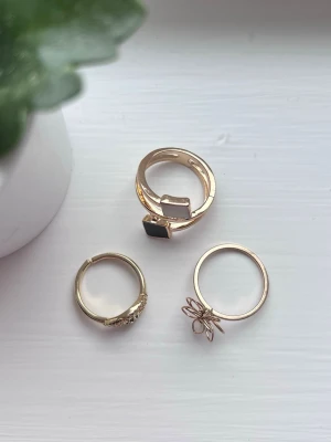 Set med tre guldfärgade ringar, mycket bra skick - Säljer ett set med tre guldfärgade ringar. En ring har två fyrkantiga stenar i svart och vitt, en har en blomformad design och den tredje har små glittrande stenar på ovansidan i form av ett öga. Blev aldrig använda. Jag lägger ringarna i plastfickan som finns på sista bilden, så de inte utsätts för skada under leverans.