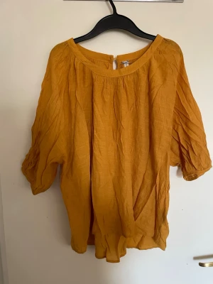Senapsgul blus med trekvartsärm - Trendig senapsgul blus i luftig bomull med avslappnad passform och rund halsringning. Blusen har trekvartsärmar med lätt puff och knappdetalj bak i nacken. Perfekt för dig som gillar en chill och stilren look.