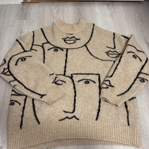 Beige stickad tröja med ansiktsmönster från Monki - Säljer en beige stickad tröja från Monki med unikt svart ansiktsmönster över hela tröjan. Tröjan har lång ärm, ribbade muddar och hög hals. Sitter lite stort så går även och ha på sig som en kort klänning. Perfekt för dig som gillar statement-plagg! Finns dock ett litet hål på baksidan.