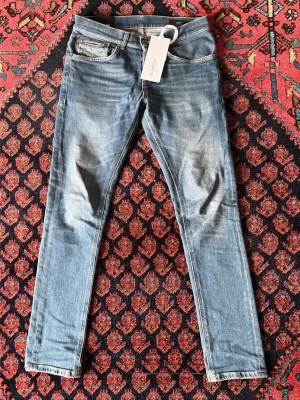 Blå slim fit jeans från dondup - Säljer ett par blå slim fit jeans med snygga slitningar och dragkedjedetaljer på bakfickorna. Har ett litet hål vid ljumsken. Jeansen har klassisk femficksdesign och normal midja. Perfekta för dig som gillar en smalare passform och en modern look.