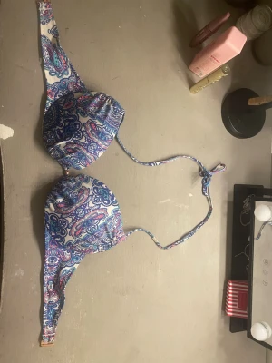 Mönstrad bikiniöverdel med paisley - Säljer en snygg bikiniöverdel med paisleymönster i blått, lila, turkost och rosa. Modellen har vadderade kupor och knytband i nacken. Materialet är troligtvis en stretchig blandning av polyester och elastan, perfekt för stranden eller poolen.