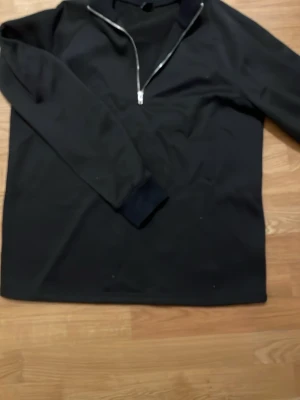 Svart halvzip tröja - En svart långärmad tröja med halvzip och hög krage. Tröjan har en enkel och clean design med muddar vid ärmsluten. Perfekt för lager på lager och funkar till det mesta. Materialet ser ut att vara mjukt och bekvämt.