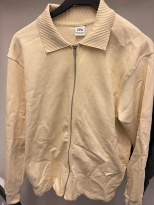 Beige ziptröja från Zara - Snygg beige ziptröja från Zara med ribbad krage och dragkedja framtill. Tröjan har en clean look och är tillverkad i ett mjukt material som passar perfekt till lager på lager. Enkel att matcha med olika outfits.