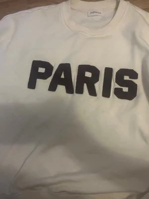 Vit sweatshirt med PARIS-tryck - Vit sweatshirt oanvänd från Even&Odd med stort svart PARIS-tryck på bröstet. Tröjan har rund halsringning och är tillverkad i mjukt material, perfekt för en avslappnad och trendig look.