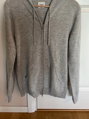 Ljusgrå hoodie från John Henric - Snygg ljusgrå hoodie från John Henric med dragkedja framtill och två fickor. Tillverkad i mjuk kashmir med ribbade muddar och justerbar huva med snören. Perfekt för en avslappnad och stilren look.