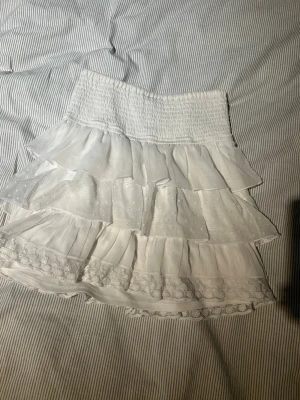 Vit volangkjol från Lindex - Supersöt vit kjol från Lindex med flera lager volanger och smock i midjan. Kjolen har fina spetsdetaljer och prickigt mönster på ett av lagren. Perfekt för dig som gillar en romantisk och luftig stil.