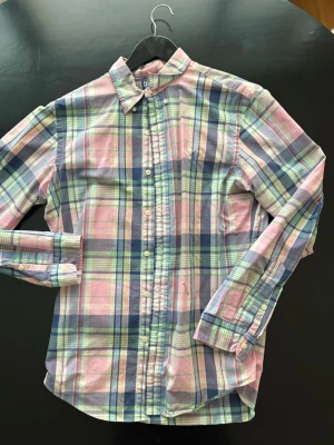 Ralph Lauren skjorta - Säljer en klassisk rutig skjorta från Ralph Lauren i pastellfärger som rosa, blå, grön och vit. Skjortan har lång ärm, knappar framtill och den ikoniska broderade loggan på bröstet. Perfekt för dig som gillar preppy stil och vill ha något färgglatt i garderoben.