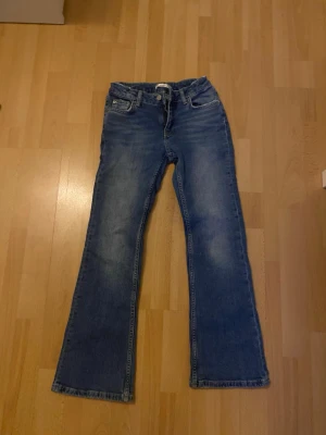 Blå bootcut jeans - Säljer ett par blå bootcut jeans med lätt slitna detaljer och klassisk femficksmodell. Jeansen har en något utsvängd passform nedtill och är tillverkade i denim. Perfekta för en avslappnad och trendig look. Storlekslappen är bortklippt men skulle gissa på storlek 152-158