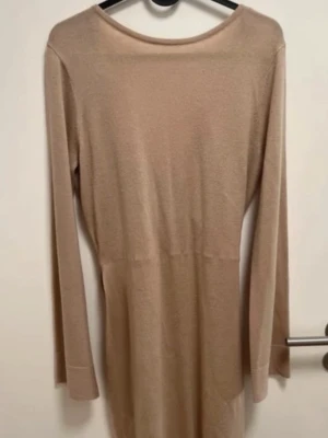 Beige omlottklänning med bälte - Stilren beige omlottklänning med lång ärm och knytband i midjan. Klänningen har en mjuk, stickad känsla och v-ringad omlottdesign baktill. Perfekt för dig som gillar enkel och elegant stil.