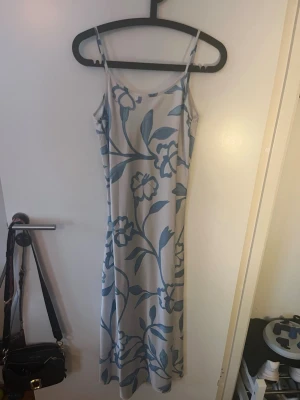 Lång blommig klänning med smala band - Ljusblå och vit långklänning med blommigt mönster och smala axelband. Klänningen har en figurnära passform och är tillverkad i ett mjukt, lätt material som passar perfekt för varma dagar. Snygg och enkel att styla med sneakers eller sandaler.