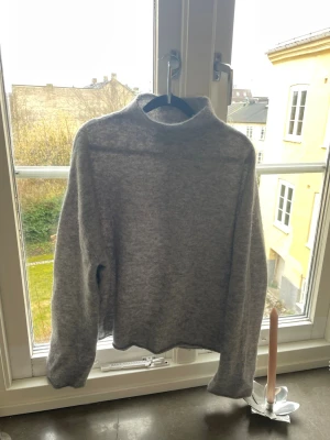 Grå stickad tröja från & Other Stories - Mysig grå stickad tröja från & Other Stories med hög hals och avslappnad passform. Tröjan har långa ärmar och är tillverkad i ett mjukt, fluffigt material som passar perfekt för kyliga dagar.