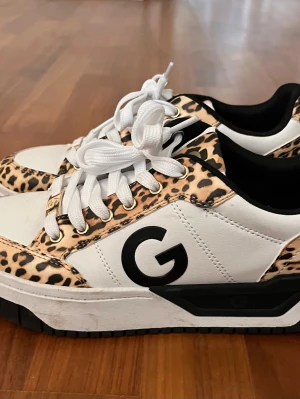 Leopardmönstrade sneakers från Guess - Snygga sneakers från Guess med leopardmönstrade detaljer, vita partier och svarta inslag. Skorna har en stor svart G-logga på sidan, vita skosnören och svart sula. Materialet är en mix av läderimitation och textil. Perfekta för dig som vill sticka ut med din stil.
