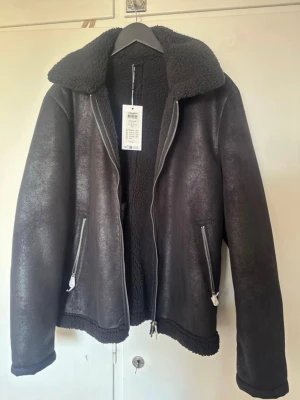 Svart teddyjacka från Jack & Jones - Snygg svart teddyjacka från Jack & Jones med mjukt, fluffigt foder och bred krage. Jackan har två dragkedjeförsedda fickor, grova silverfärgade dragkedjor och en klassisk rak passform. Perfekt för kalla dagar och ger en cool look.