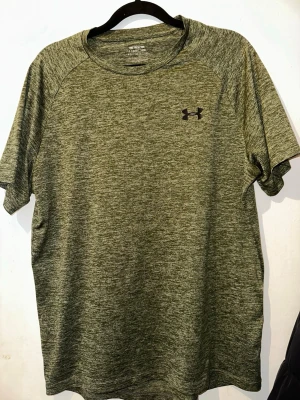 Grön tränings-tshirt från Under Armour - Snygg grönmelerad tränings-tshirt från Under Armour med klassisk logga på bröstet. Tillverkad i ett lätt och snabbtorkande funktionsmaterial som passar perfekt till gymmet eller löprundan. Rund halsringning och korta ärmar för maximal rörelsefrihet.