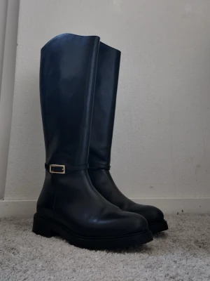 Svarta höga stövlar Stockholm Design Group - Snygga svarta höga stövlar från Stockholm Design Group i slätt läder. Stövlarna har en chunky sula och en liten gulddetalj vid ankeln. Perfekta för att lyfta vilken outfit som helst och ger en clean, modern vibe.